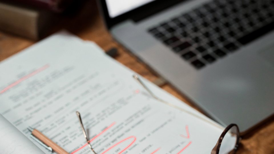 A manuscript covered in red editorial marks on a desk beside reading glasses and a laptop.
