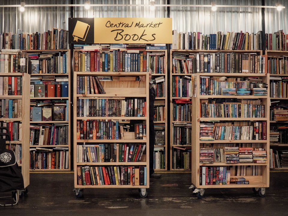 Wheeled shelving units in a market bookshop, packed with second-hand books under a hand-lettered sign.