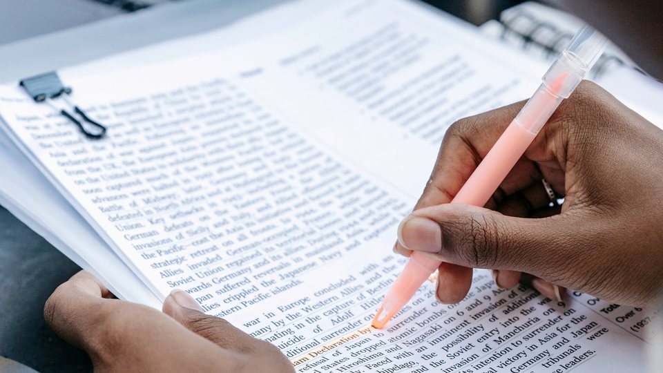 A hand running a highlighter across a page of a printed manuscript, the stack clipped together.