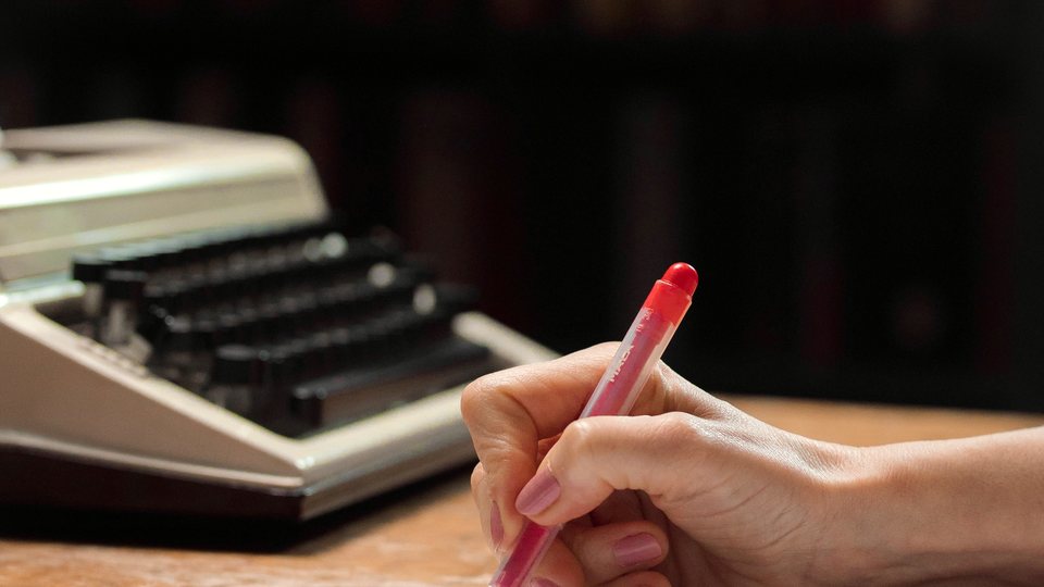 A hand holding a red editorial pencil over a desk, an old typewriter beside it.